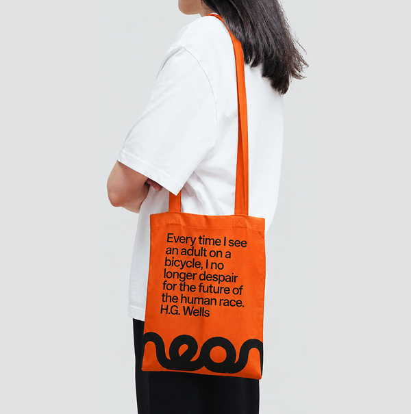 The neon tote: orange canvas with an H.G. Wells quote and the neon wordmark, worn over the shoulder